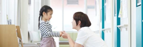 女性が廊下でひざまずき、ストライプのシャツとピンクのスカートを着た少女と手をつないでいる。.