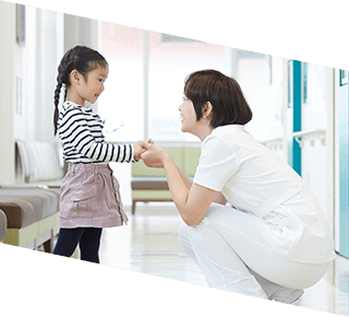 看護師が廊下でひざまずいて少女と手を繋いでいる。少女はストライプのシャツとスカートを着ている。.