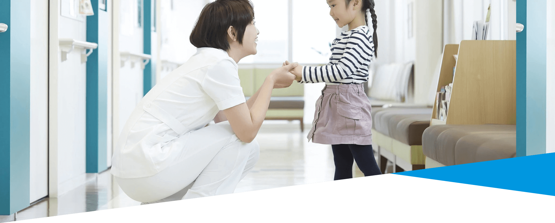 看護師が廊下で少女とひざまずいて手を握り、お互いに微笑んでいる。少女はストライプのシャツと紫のスカートを着ている。.