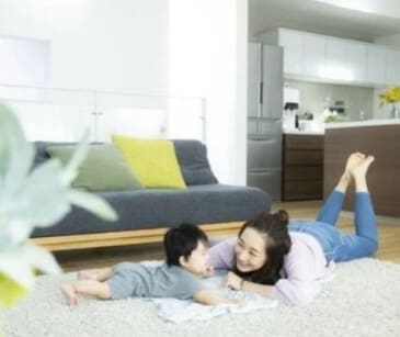 女性と赤ちゃんが、ソファとキッチンを背景にしたリビングルームのカーペットの上に横たわっています。.