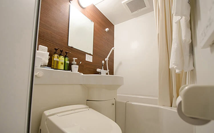 トイレ、洗面台、鏡、シャワーと浴槽が一体となった小さなバスルーム。洗面台にはアメニティのボトルが置いてあります。茶色のアクセント壁とベージュのシャワーカーテン。頭上には天井ライトがあります。.