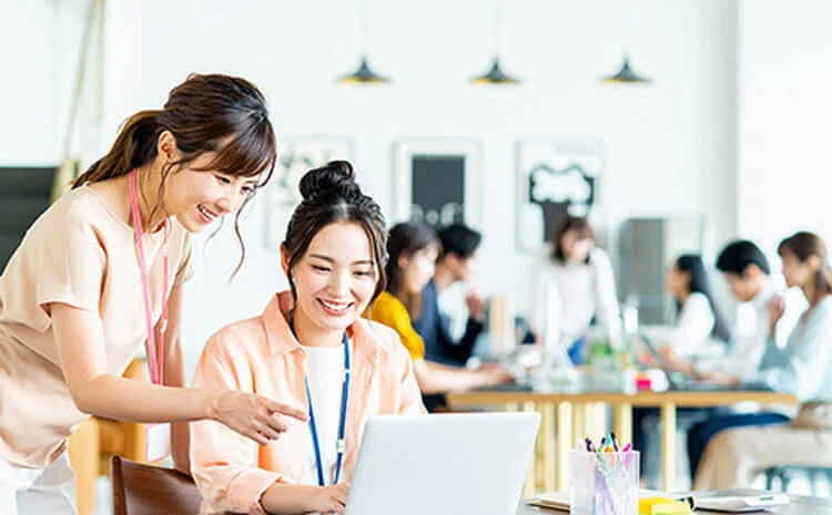 忙しいオフィスで、2 人の女性がノートパソコンを見ながら微笑んでいます。背景では他にも数人が働いています。.