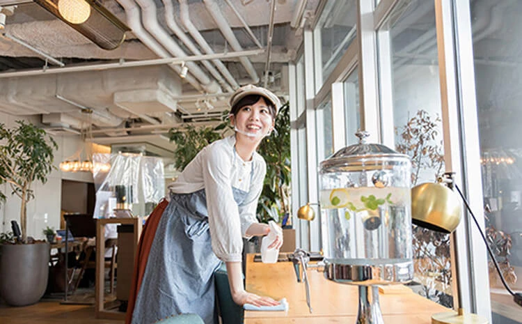 白いシャツとエプロンを着た女性が、モダンなカフェの大きな窓の近くのテーブルを掃除しています。背景にはガラスのウォーターディスペンサーと緑の植物があります。.