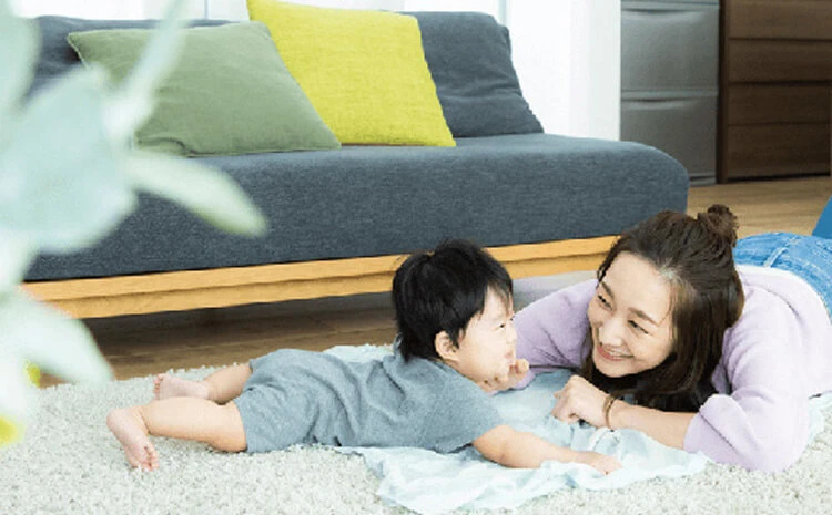 室内の敷物の上に横たわり、向かい合って微笑んでいる女性と赤ちゃん。背景にはソファがある。.