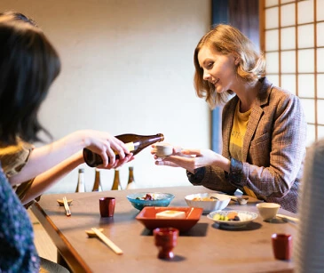 2 人の人が日本食レストランのテーブルに座っています。テーブルにはさまざまな料理が置かれており、1 人がもう 1 人に飲み物を注いでいます。.