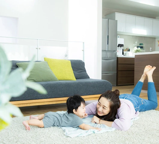 明るいリビングルームで、柔らかいラグの上に赤ちゃんを置き、床に横たわる女性。背景にはソファとキッチンがあります。.