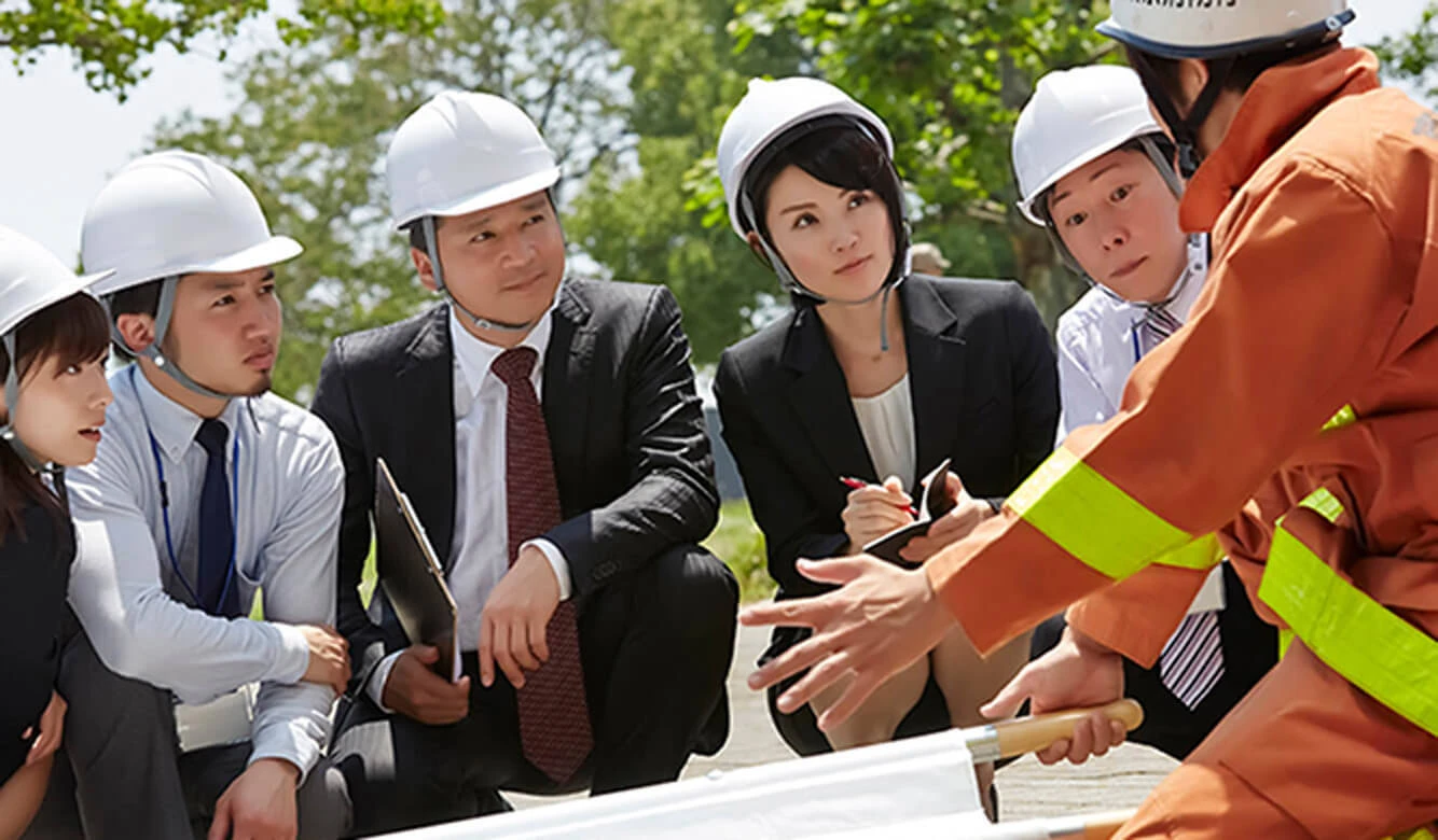 ヘルメットとビジネス服を着た人々のグループが、屋外で身振り手振りを交えながら何かを説明しているオレンジ色の制服を着た人の話を注意深く聞いています。.