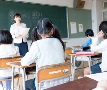 教室の風景。生徒たちは机に座り、緑の黒板のそばの先生と向き合っています。生徒の中には書いている子もいれば、聞いている子もいます。先生は紙を持って話しています。.