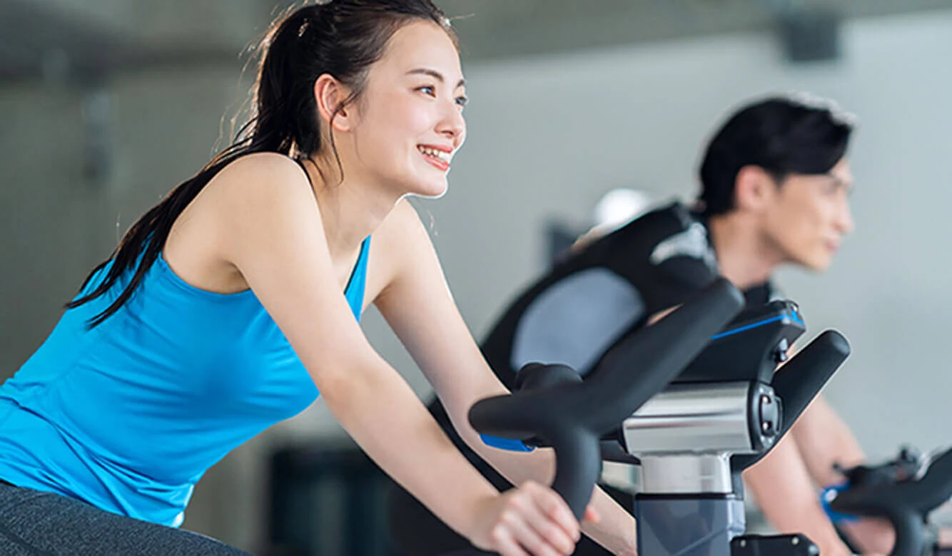 2 人がジムでエアロバイクで運動しており、1 人は青いタンクトップを着て笑顔です。.