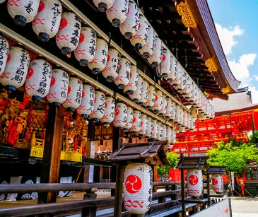 赤い木造建築と澄んだ青い空を背景に、伝統的な日本寺院の外に、日本語の文字が書かれた白い提灯が一列に吊るされています。.