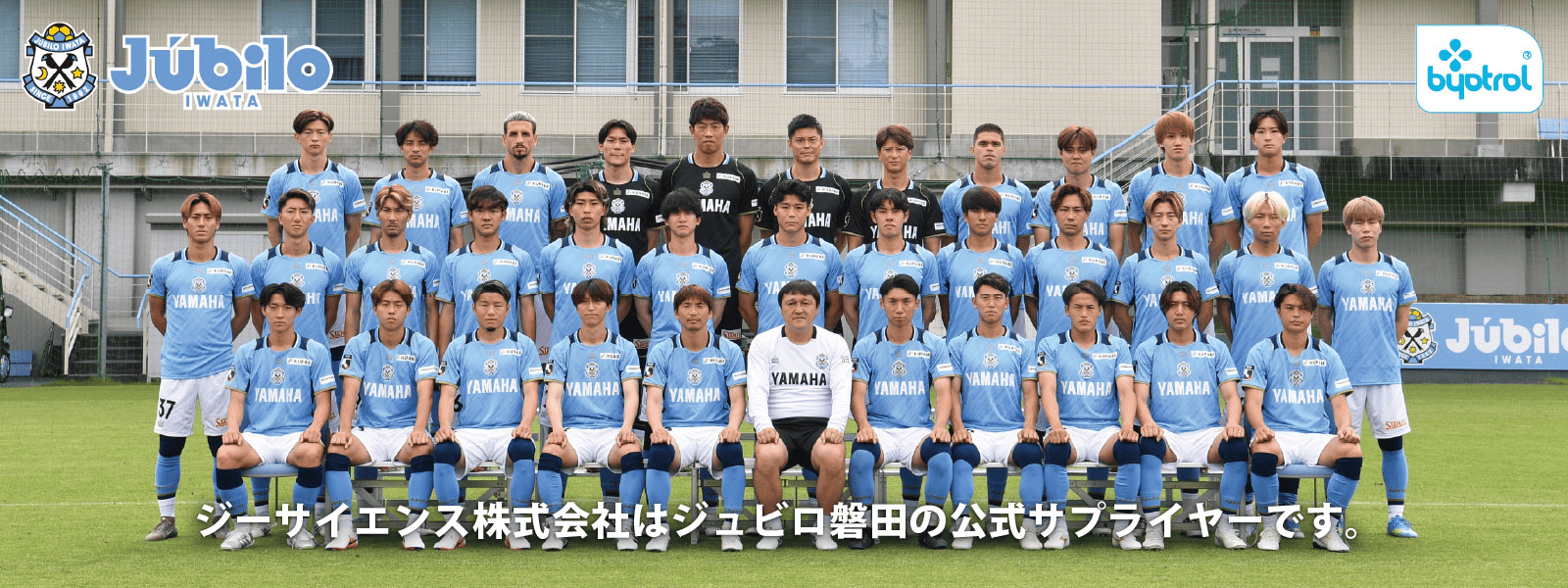 青と白のユニフォームを着たサッカーチームが、ジュビロ磐田（Júbilo Iwata）のロゴやスポンサーのバナーが飾られた建物の前で、コーチやスタッフと共に芝生の上で集合写真を撮影している。この光景は、同チームがJ2リーグに所属していることや、公式サプライヤーとのつながりを際立たせている。.