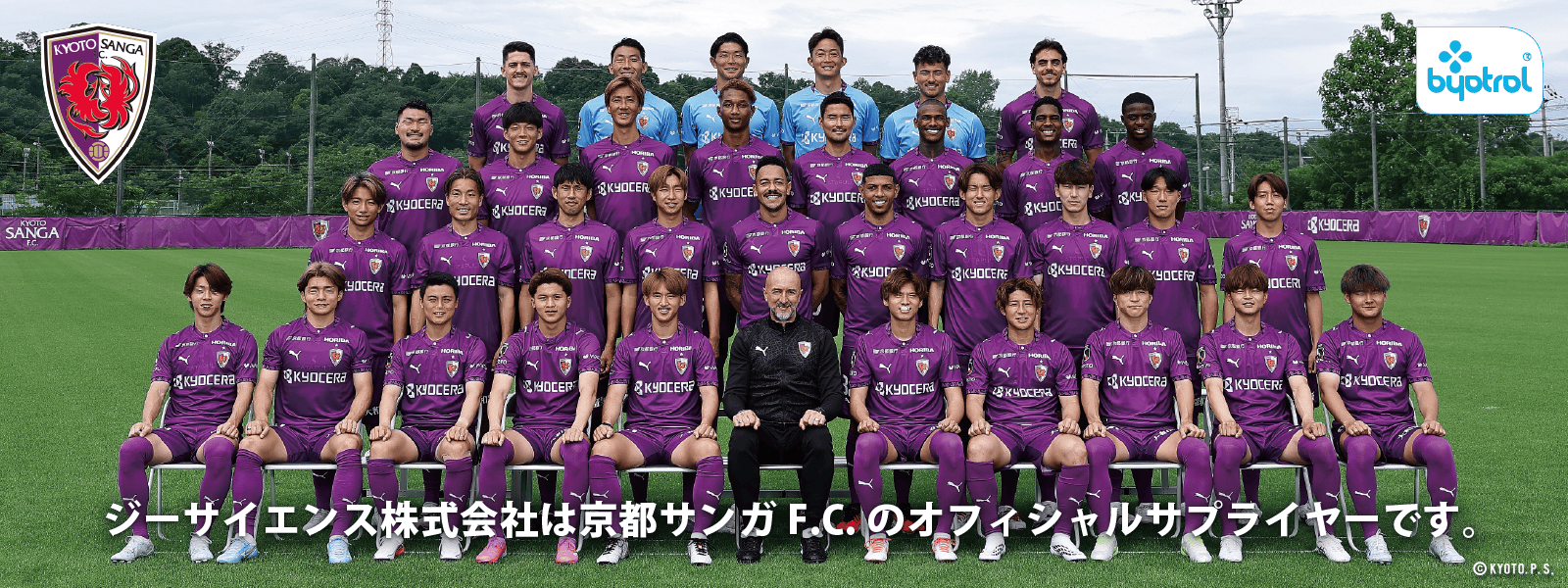 J1リーグに所属する誇り高きクラブ、京都サンガF.C.のサッカーチームが、木々やグラウンドを背景に、コーチやスタッフと共に、紫色のユニフォーム姿で屋外で集合写真を撮影した。この写真は、オフィシャルサプライヤーの支援を受けて撮影されたものである。.